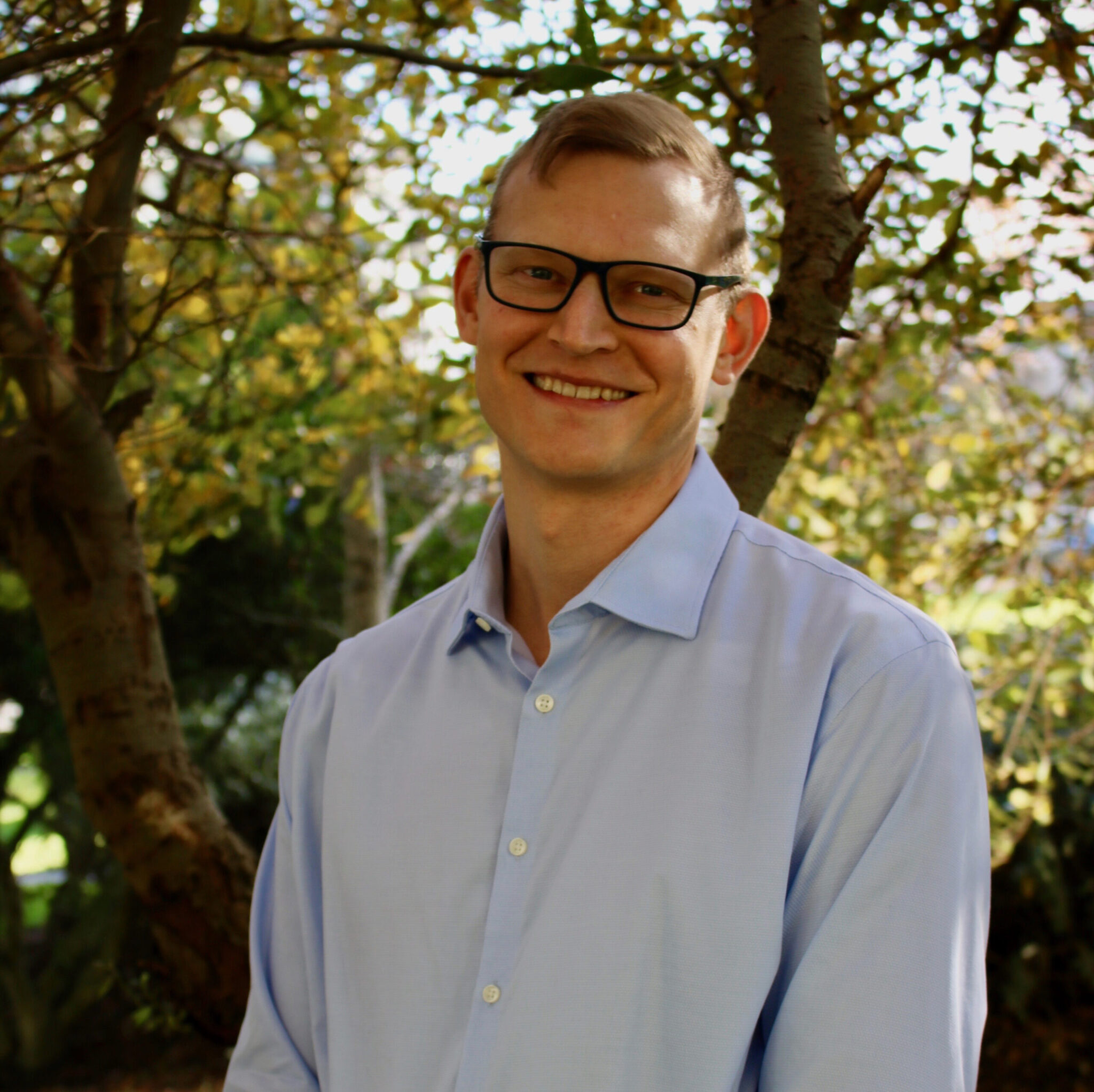 Photo of a smiling white man wearing thick-rimmed glasses and a light blue dress shirt.