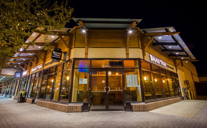 Nighttime photo looking into a warmly lit restaurant with glass walls and a door on the corner. Sign says Baan Thai