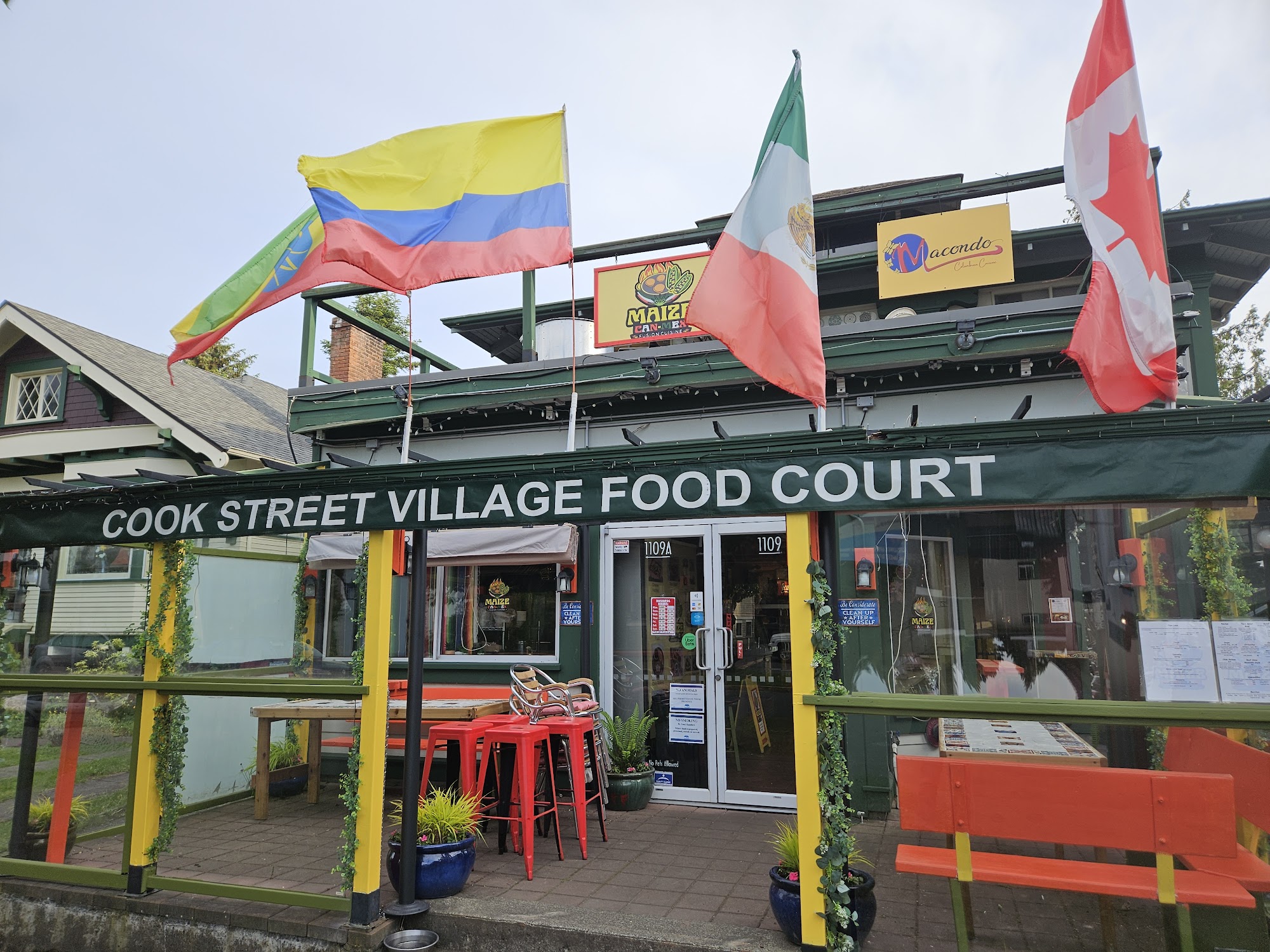 Photo of a patio with colourful tables and chairs, with a banner that reads Cook Street Village Food Court