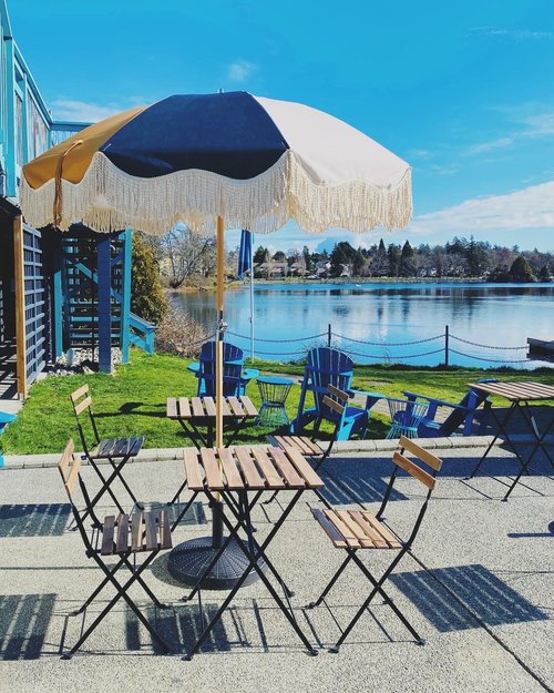 Photo of an outdoor bistro table with 2 chair and a fringed umbrella, with ocean in the background