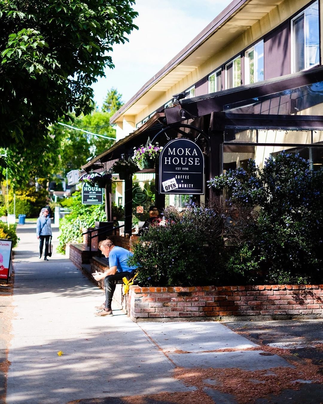 Photo of a covered, raised patio along a sidewalk, with greenery and tall trees.